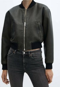 Chaqueta bomber corta de satén verde oscuro con cierre de cremallera, puños acanalados y bolsillos laterales, combinada con jeans negros.