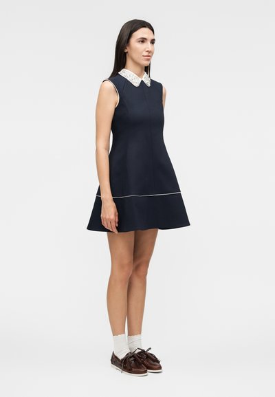 Γυναίκα στέκεται φορώντας αμάνικο φόρεμα σε ναυτικό μπλε με λευκό δαντελένιο γιακά, λευκές κάλτσες και καφέ δερμάτινα παπούτσια τύπου boat shoes σε ουδέτερο φόντο.