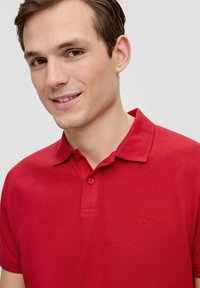 Rotes Poloshirt aus Baumwolle. Merkmale sind ein Kragen, eine Knopfleiste mit zwei Knöpfen und ein strukturiertes Gewebe. Gesticktes Logo auf der linken Seite.