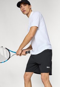 Bela polo majica z ovratnikom in gumbi, kombinirana s črnimi športnimi hlačami z logotipom. Model drži teniški lopar z modrimi poudarki.