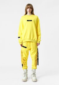 Sudadera amarilla y pantalones a juego con etiquetas negras, patrones texturizados únicos en los pantalones y botas blancas con cordones y suelas gruesas.