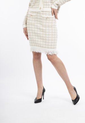 Gewebter cremefarbener Rock mit strukturiertem Muster, Fransen-Saum und zwei vorderen Taschen; kombiniert mit schwarzen High-Heels.