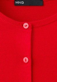 Cardigan rouge en maille avec un col rond et deux boutons rouges visibles, montrant une étiquette noire avec le logo blanc MNG.