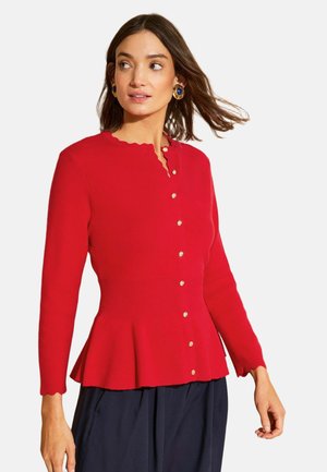 Žena oblečená v červenom kardigáne so zapínaním na gombíky a vlnitými okrajmi a tmavomodrými nohavicami, otočená hlavou do strany, s vlasmi jemne vejárovo pofukovanými.