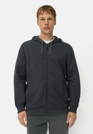 Junger Mann trägt einen dunklen Kapuzenpullover mit Reißverschluss und Kordeln, steht mit locker an den Seiten hängenden Händen vor einem schlichten hellen Hintergrund.