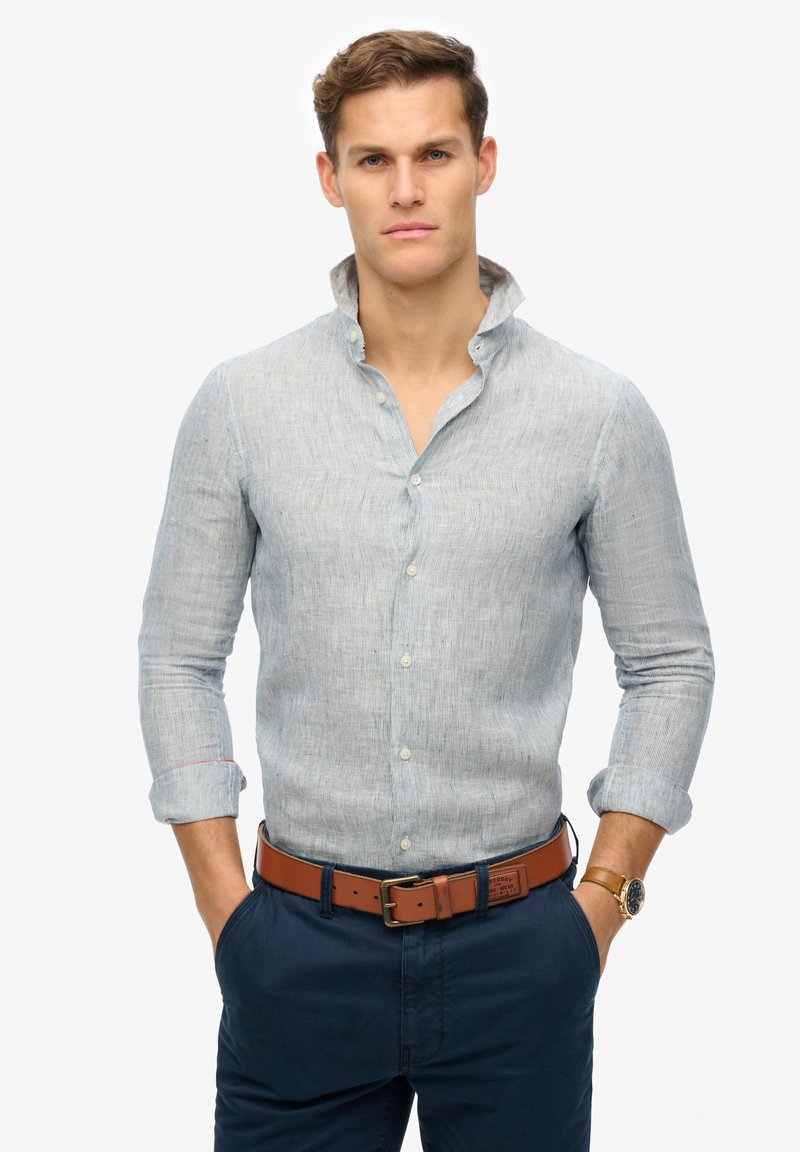 Camicia button-up di lino azzurro chiaro con colletto alla coreana, maniche lunghe arrotolate ai polsi, abbinata a pantaloni blu navy e una cintura marrone.