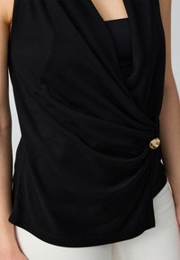 Top nero senza maniche con frontale drappeggiato e design attorcigliato, dettaglio in bottone dorato e tessuto liscio ed elastico. Vestibilità su misura.