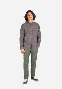 Young man with shoulder-length hair wearing a brown button-up shirt, green pants, and gray sneakers standing with one hand in pocket.