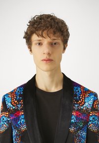 Herreblazer med fargerike sommerfuglmønstre i blå, oransje og rød på stoffet; svarte satengklipper; brukt over en svart skjorte.