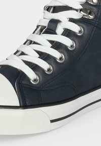 Σκούρο μπλε ψηλοτάκουνο αθλητικό παπούτσι με λευκό καπάκι από λάστιχο στα δάχτυλα, λευκά κορδόνια περασμένα από μεταλλικά ροδέλες και λευκή σόλα με μαύρη ρίγα.