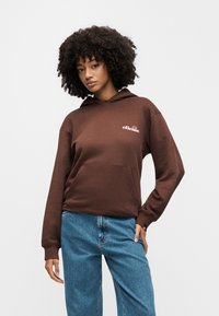 Jeune femme aux cheveux bouclés portant un sweat à capuche marron de la marque ellesse et un jean bleu, posant avec une main sur la hanche sur un fond blanc.