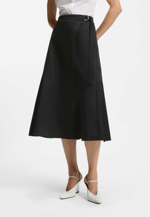 Kobieta ubrana w czarną midi spódnicę kopertową z paskiem, białą bezrękawnikową bluzkę oraz białe sandały na obcasie z paskiem na pięcie, stojąca.