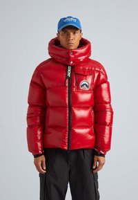 Giacca a vento rossa con finitura lucida, colletto alto e zip frontale. Presenta una patch con il logo sul petto, indossata sopra pantaloni neri.