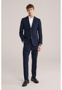 Moški v mornarsko modrem suknjiču z belo srajco in črnimi poslovnimi čevlji, stoji z eno roko v žepu na nevtralni podlagi.