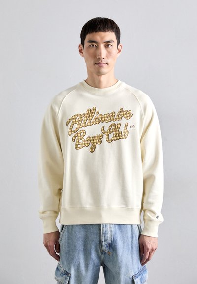 Homme portant un sweat-shirt crème avec le texte "Billionaire Boys Club" dans un motif de grille marron, associé à un jean cargo bleu clair, se tenant devant un mur uni.