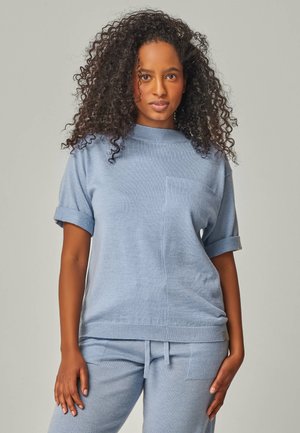 Hellblauer Strickpullover mit kurzen, umgeschlagenen Ärmeln und einer Brusttasche, kombiniert mit passenden Hosen. Weiche Textur und lockere Passform.