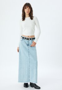 Witte langemouwen cropped top gecombineerd met een lichtblauwe denim maxi rok met een voorwaartse split en een rafelige zoom, aangevuld met een zwarte riem.
