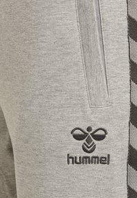 Graue Sweatpants mit einem schwarzen gestickten Logo, Seitentaschen und einem strukturierten Design entlang der äußeren Naht im Zickzackmuster.