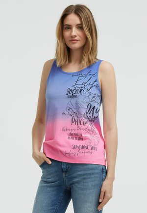 Frau mit schulterlangem Haar, die ein ärmelloses Top mit blau-rosa Farbverlauf, schwarzem Text und grafischen Designs trägt, kombiniert mit blauen Jeans.