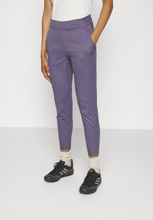 Person iført lilla tætsiddende joggebukser, beige krøllede sokker, sorte sneakers og en hvid top, stående med den ene hånd i lommen.