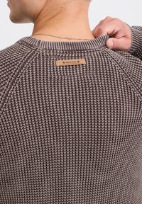 Sweter dzianinowy w ciemnym brązie, z teksturowanym wzorem, żebrowanym kołnierzem oraz skórzaną metką z napisem "INDICODE" na plecach.