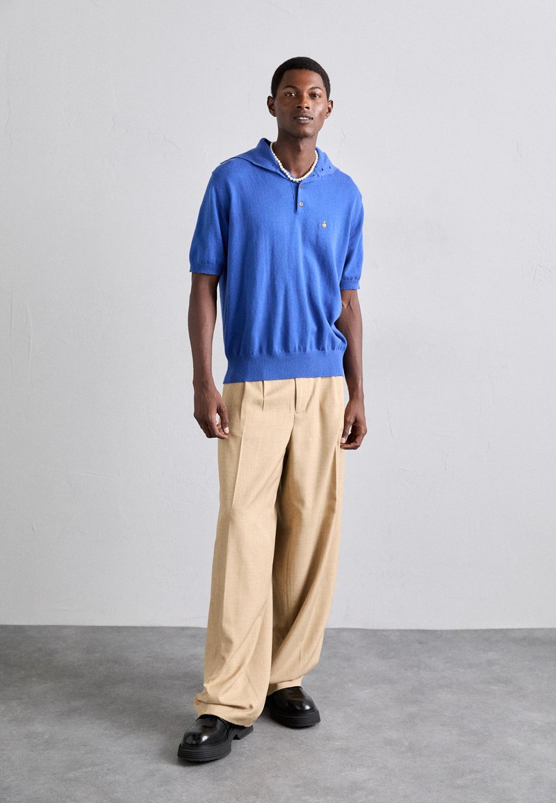 Blauwe gebreide polo met korte mouwen en een capuchon, voorzien van een sluiting met knopen aan de voorkant; gecombineerd met beige wijde broekspijpen en zwarte chunky schoenen.