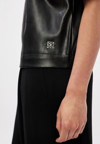 Top de couro preto com uma textura suave, apresentando um detalhe em logótipo prateado perto da bainha. Combinado com calças pretas ajustadas.