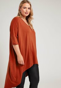 Rostfärgad, löst sittande topp med djup V-hals, korta ärmar och asymmetrisk fåll. Tillverkad av mjukt, lätt material.