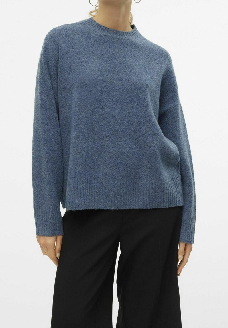Femme portant un pull en laine doux bleu à manches longues et un pantalon noir, avec des boucles d’oreilles anneaux et les mains détendues le long du corps.