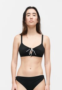 Mujer de pie mirando hacia adelante, vistiendo un bikini negro con un detalle de lazo blanco en la parte superior sobre un fondo blanco liso.