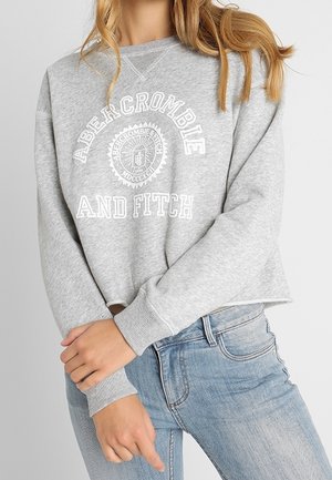 Femme portant un sweat-shirt gris clair Abercrombie and Fitch et un jean bleu, posant bras croisés à la taille.