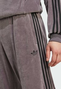 Zatvoren kadar osobe koja nosi sive Adidas trenirke i jaknu s crnim trostrukim detaljima i logom na hlačama. Ruka opuštena uz tijelo.