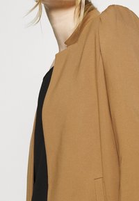 Hnědý blazer s hladkým povrchem, vyznačující se stojáčkem a jemným nařasením na ramenou, který je doplněn černým topem pod ním.