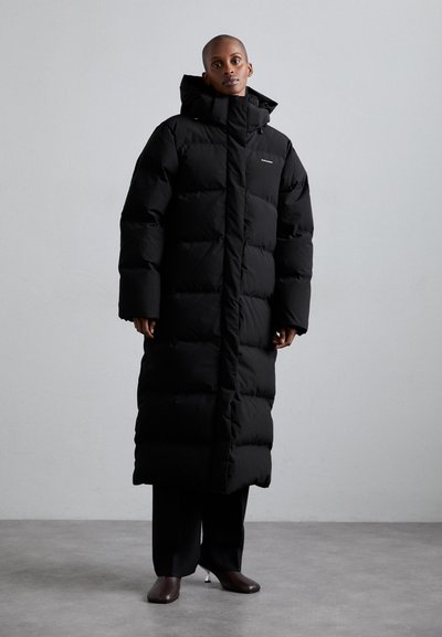 Cappotto lungo nero in piumino con cappuccio, caratterizzato da un design trapuntato, colletto alto e chiusura a bottone nascosta. Struttura liscia e vestibilità oversize.