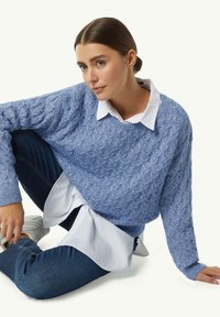 Gelaagde textuur blauwe gebreide trui over een witte blouse met knopen, met een ronde halslijn en geribbelde manchetten, gedragen met donkere spijkerbroek.
