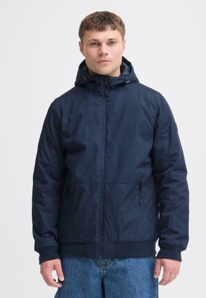 Jeune homme portant une veste à capuche bleu marine et un jean bleu, debout devant un fond gris clair uni.