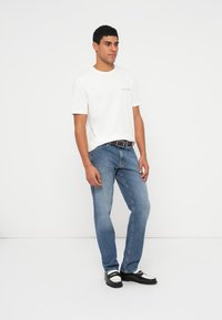 Witte katoenen T-shirt gecombineerd met lichtblauwe spijkerbroek. Zwarte riem en zwart-witte schoenen maken de outfit compleet. Staande model, neutrale achtergrond.