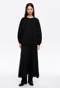 Sudadera negra de manga larga con detalles de tachuelas en los hombros, combinada con una falda maxi negra con pliegues y botas negras hasta el tobillo.