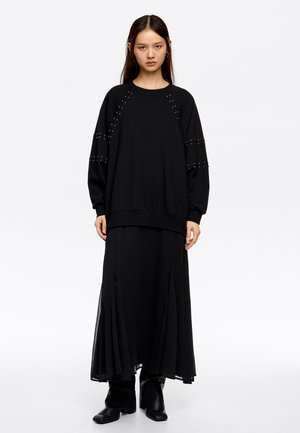 Sudadera negra de manga larga con detalles de tachuelas en los hombros, combinada con una falda maxi negra con pliegues y botas negras hasta el tobillo.