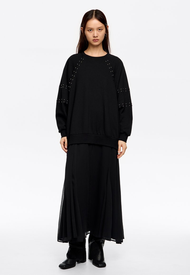 Sudadera negra de manga larga con detalles de tachuelas en los hombros, combinada con una falda maxi negra con pliegues y botas negras hasta el tobillo.