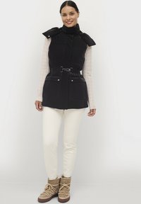 Gilet nero in piuma con vita cinturata, colletto alto, bottoni a scatto e due tasche frontali, indossato sopra una maglietta a maniche lunghe con fantasia e pantaloni crema.