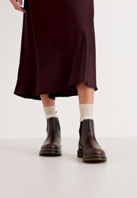 Stivali ankle in pelle marrone con punta rotonda, suola nera e cuciture verdi, indossati con calze a maglia color crema e una gonna bordeaux scura.