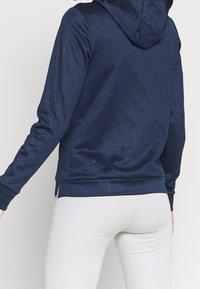 Mörkblå hoodie med geometrisk vadderad textur, utrustad med dragsko i huvan och åtsittande mudd. Ljusreflekterande detaljer synliga.