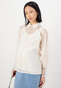 Lichtcrème blouse met ingewikkelde kantdetails, met knoopsluiting aan de voorkant en lange mouwen. Gladde stof met doorzichtig kant op de schouders en manchetten.