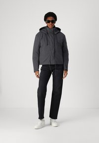 Gray padded jacket with a hood, drawstrings, and front zipper, paired with dark jeans and white sneakers. Model wears black sunglasses.