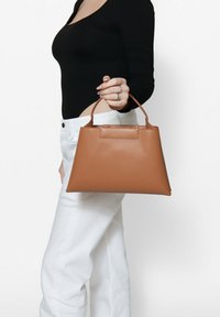 Borsa a tracolla in pelle color tan con una silhouette strutturata, manici corti e una base piatta, tenuta da una persona che indossa un top nero e pantaloni bianchi.