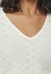 Pull en tricot blanc avec un col en V, présentant un motif texturé de cercles en relief, en matériau léger et avec des manches courtes.