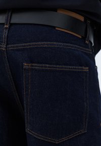 Temno modre kavbojke iz jeansa z oranžnimi šivi in enim zadnjim žepom. Vključuje črni usnjeni pas z rjavim usnjenim poudarkom.