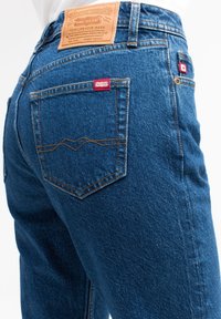 Jeans de mezclilla azul con un parche de cuero de la marca. Las características incluyen costura doble, una etiqueta roja en el bolsillo trasero y un corte clásico.