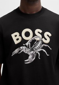 Svart bomullströja med en stor grå hummergrafik och ordet "BOSS" i beige, fet versal ovanför designen.